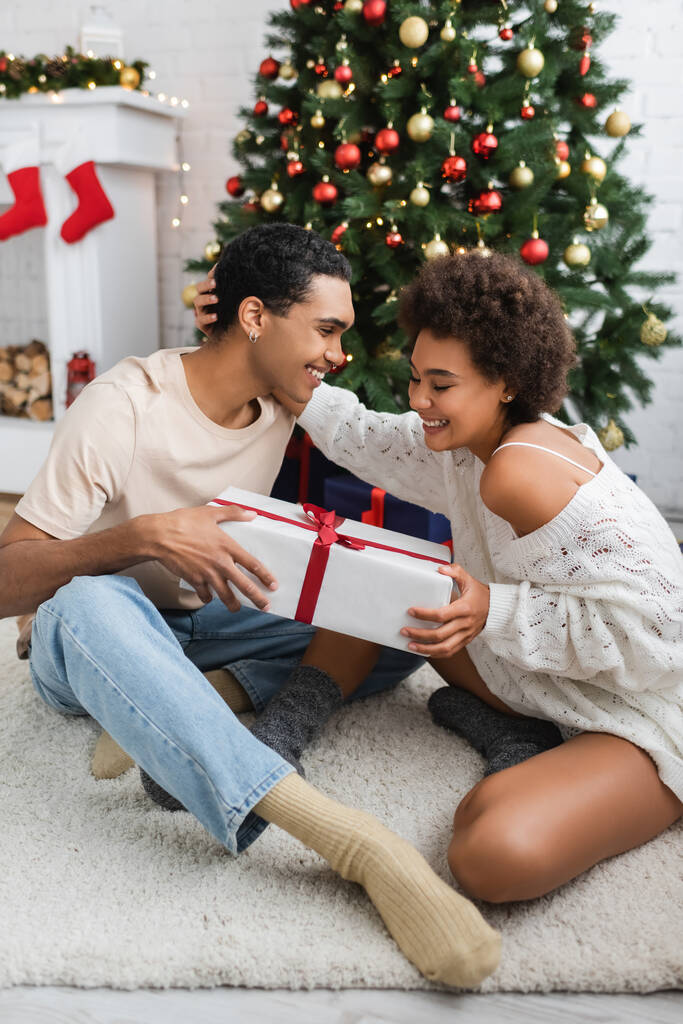 vrolijk Afrikaans amerikaans paar zitten op de vloer met geschenk doos in de buurt van kerstboom - Foto, afbeelding