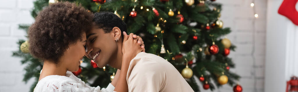 giovane donna afroamericana che abbraccia sorridente fidanzato vicino all'albero di Natale, striscione - Foto, immagini