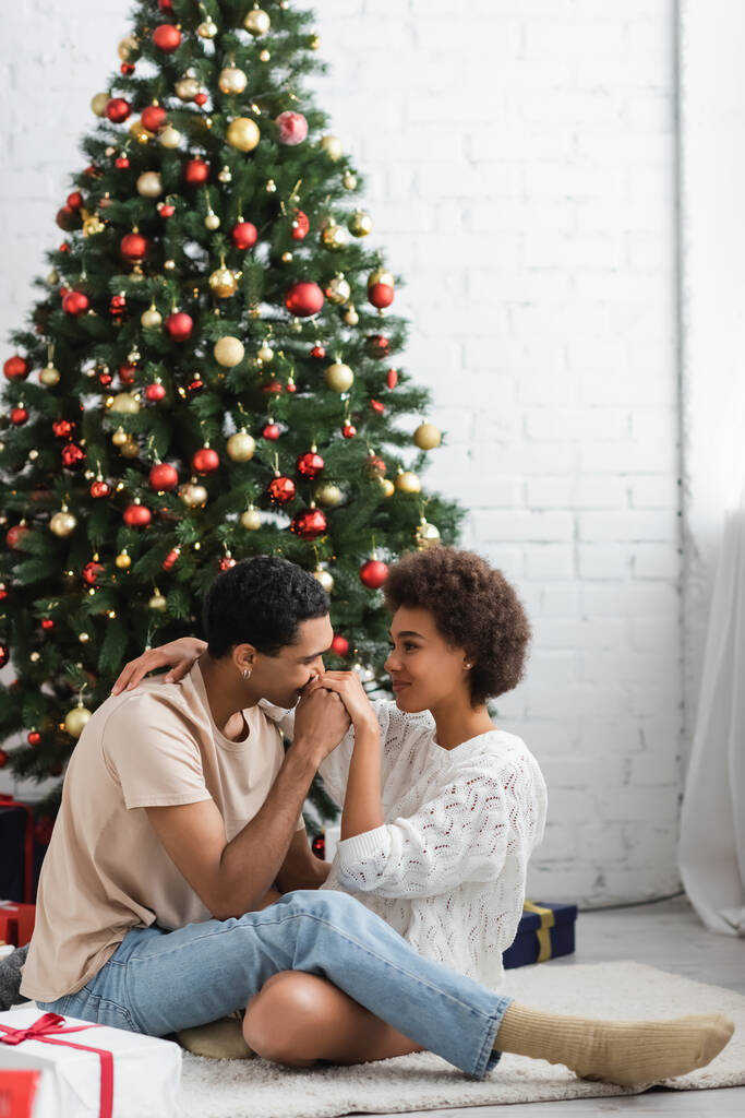 νεαρός Αφροαμερικάνος που φιλάει το χέρι μιας σέξι γυναίκας που κάθεται στο πάτωμα κοντά στο χριστουγεννιάτικο δέντρο. - Φωτογραφία, εικόνα