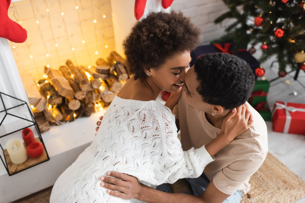 sexy african american woman in white openwork sweater embracing with man near fireplace - Photo, Image