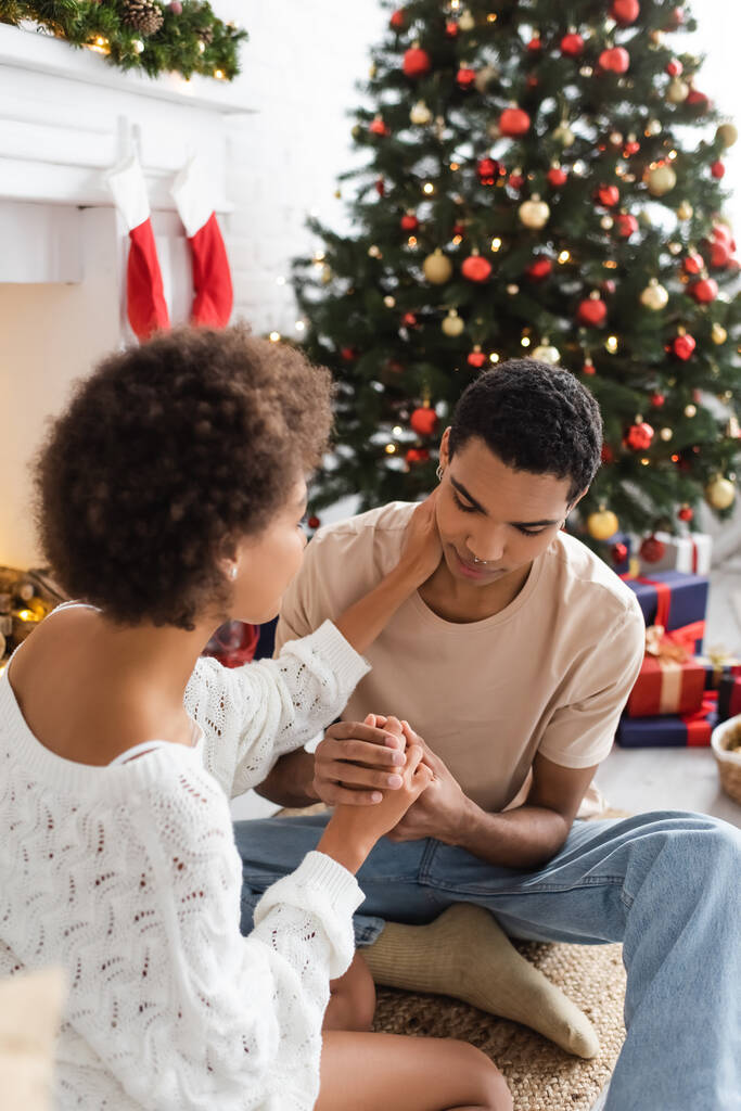 Genç Afro-Amerikan erkek kız arkadaşının elini tutuyor. Bulanık Noel ağacının yanında boynuna dokunuyor. - Fotoğraf, Görsel