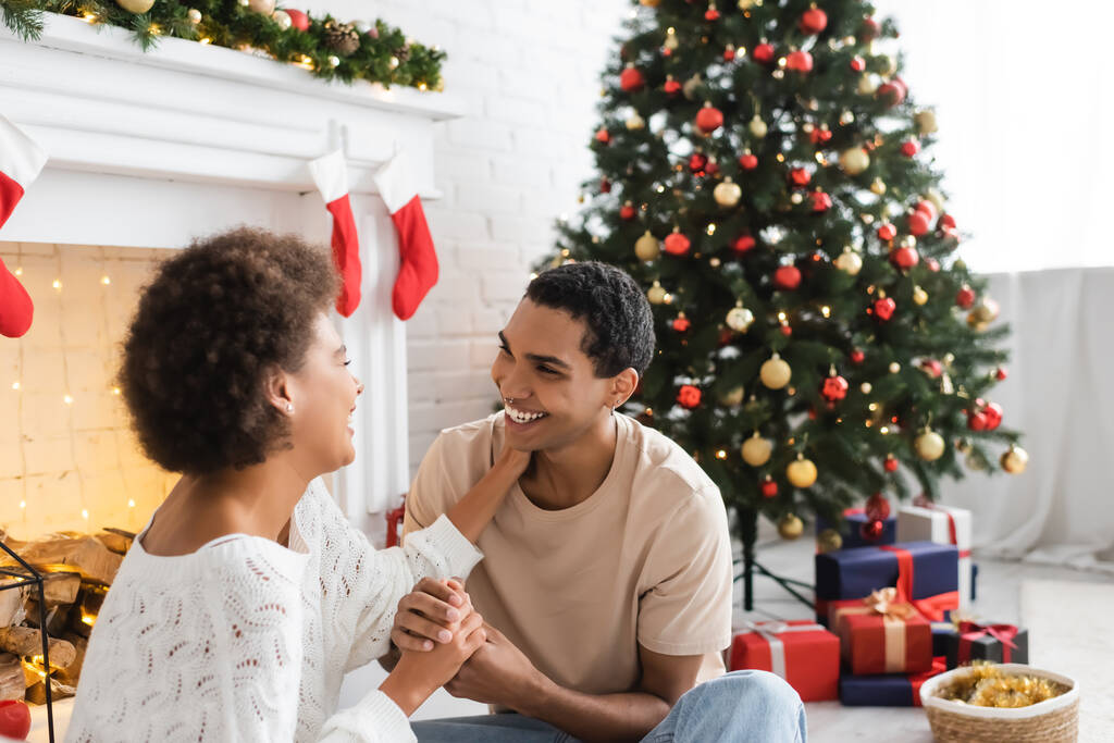 fröhliches afrikanisch-amerikanisches Paar hält Händchen und schaut sich in der Nähe von Weihnachtsbaum und Kamin an - Foto, Bild