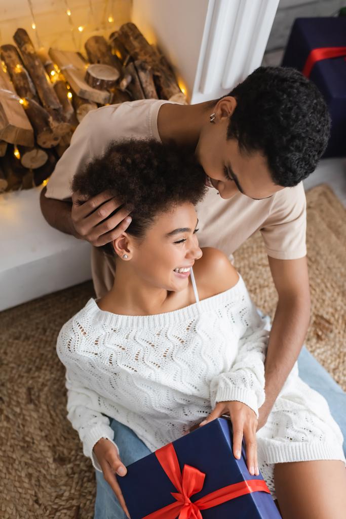 fiatal afro-amerikai férfi megérinti göndör haj boldog barátnő ül ajándékdobozzal közel kandalló - Fotó, kép