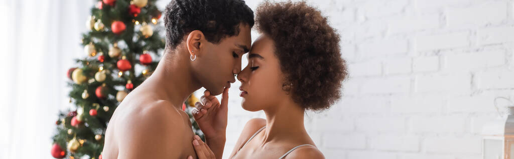 vue latérale d'une femme afro-américaine frisée touchant les lèvres d'un petit ami sexy près d'un arbre de Noël flou, bannière - Photo, image