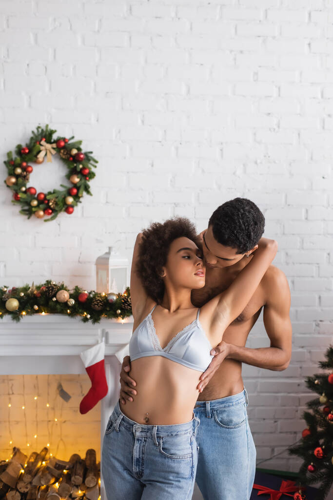 jovem afro-americano homem abraçando namorada em sutiã e jeans perto de lareira com decoração de Natal - Foto, Imagem