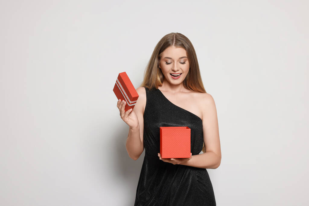 Emotionale junge Frau in elegantem schwarzen Kleid öffnet rote Geschenkschachtel auf weißem Hintergrund - Foto, Bild