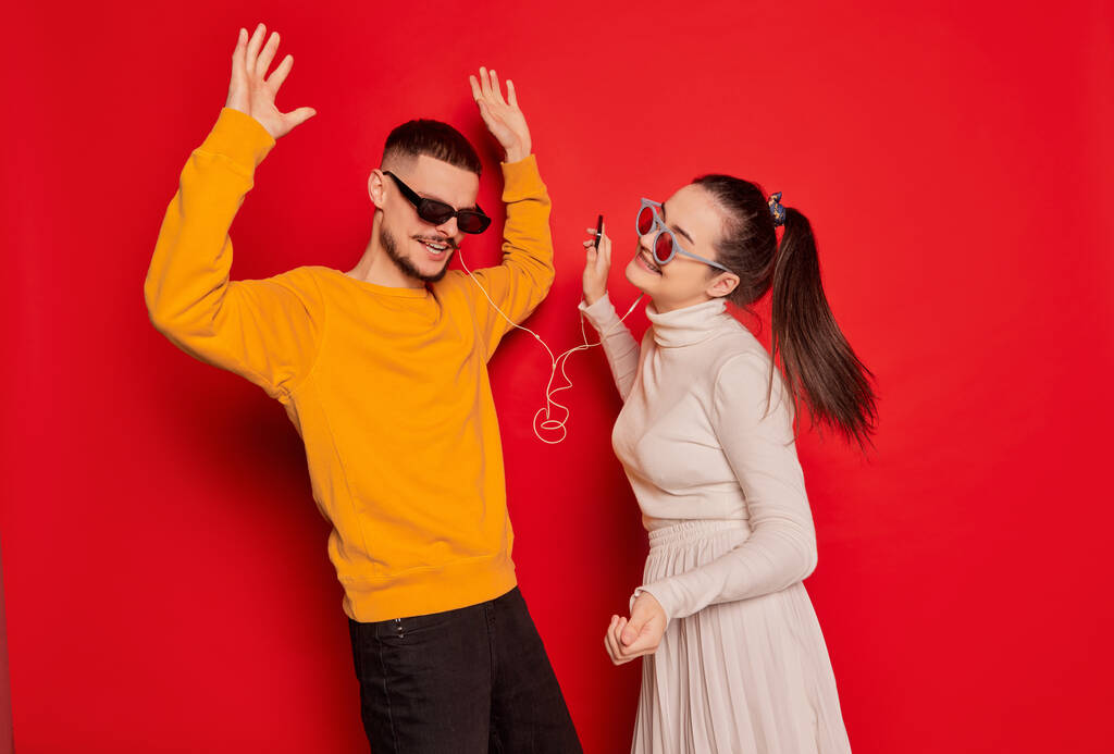 Портрет щасливої молодої пари, чоловіка і жінки, весело танцює з музикою в навушниках ізольованих на червоному фоні. Концепція любові, стосунків, Валентинів день, емоцій, способу життя, вечірки, розваг - Фото, зображення