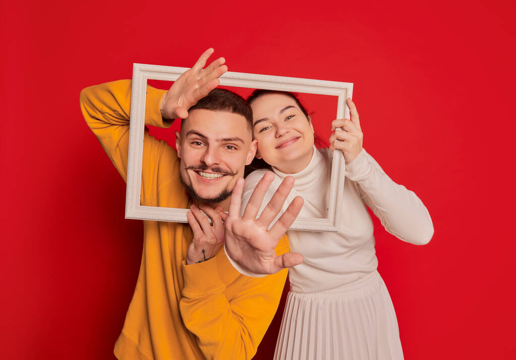 Portrait of happy young couple, man and woman, posing with picture frame isolated over red background. Looking very happy. Concept of love, relationship, Valentines Day, emotions, lifestyle - Photo, Image