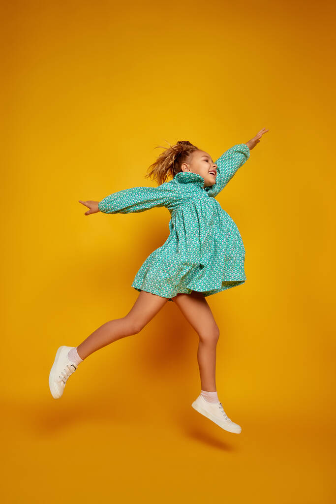 Have a fun. Happy smiling little girl, kid in color dresss jumping high isolated over yellow studio background. Kids fashion, emotions, carefree childhood, challenges, education and freedom concept - Photo, Image