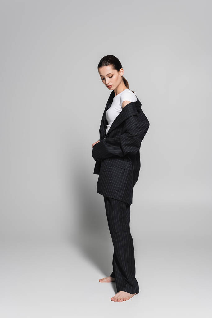 Ξυπόλητο μοντέλο σε μαύρο κοστούμι στέκεται σε γκρι φόντο με σκιά  - Φωτογραφία, εικόνα