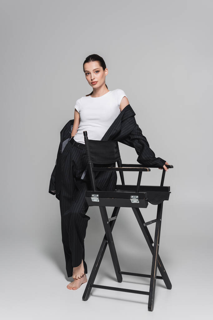 Stylish barefoot woman in black suit and t-shirt posing near folding chair on grey background  - Photo, Image