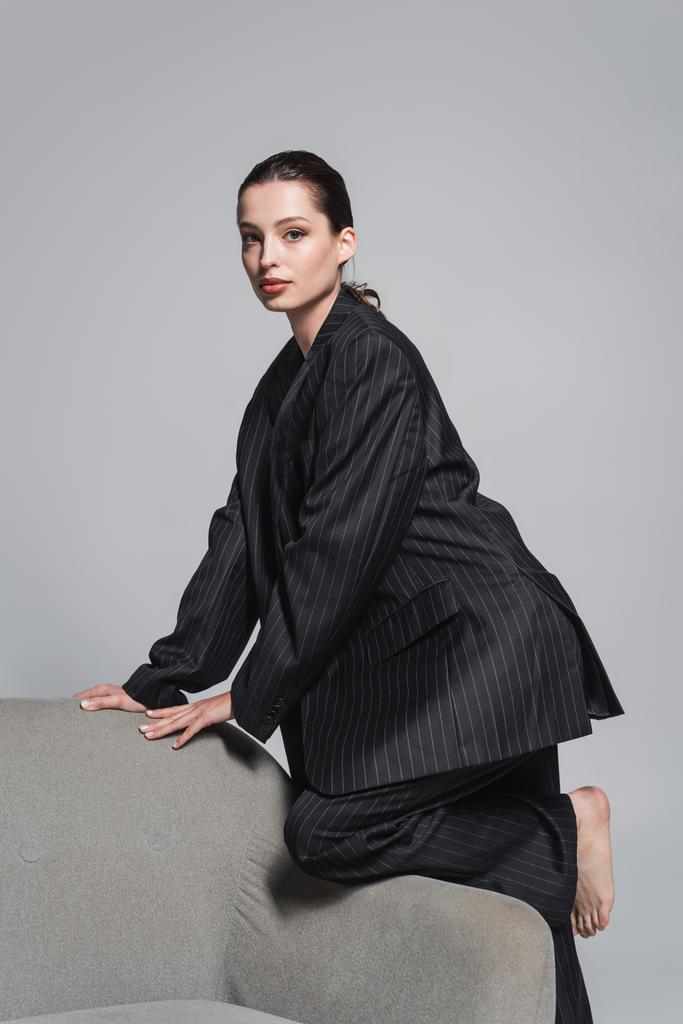 Brunette nainen raidallinen puku poseeraa lähellä nojatuoli eristetty harmaa  - Valokuva, kuva