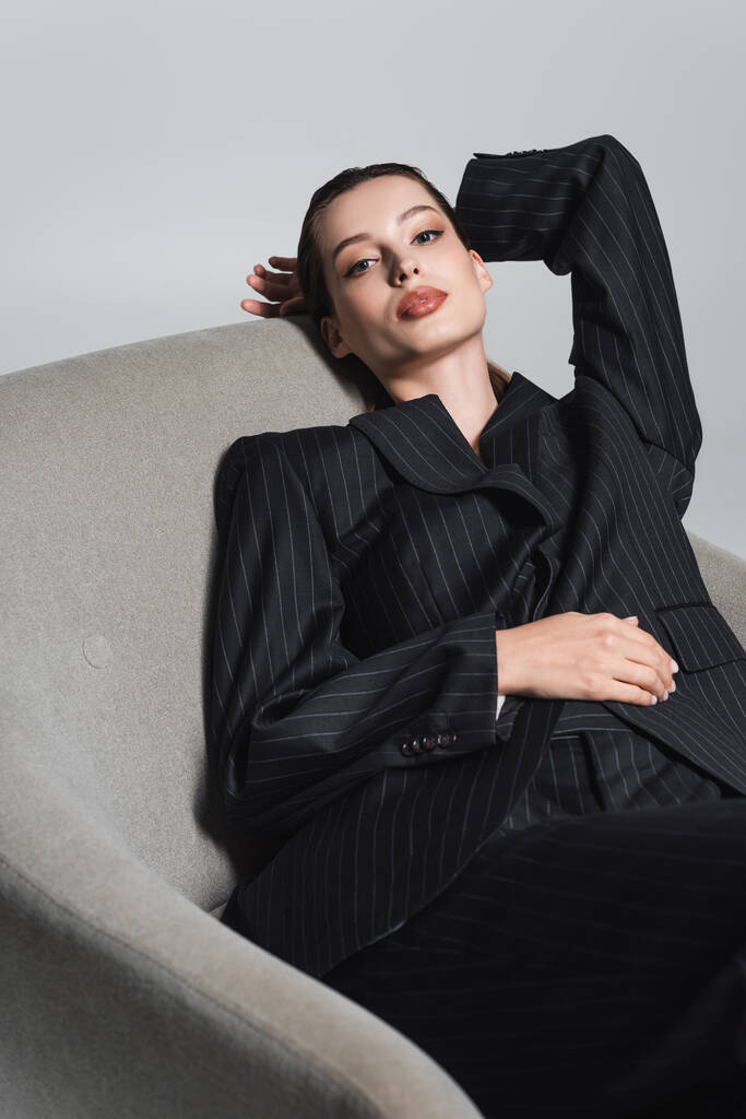 Stylish woman in black suit posing on armchair isolated on grey  - Photo, Image