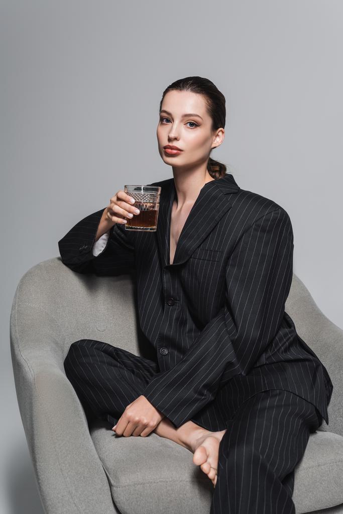 Modelo elegante em terno preto segurando vidro de uísque enquanto sentado em poltrona em fundo cinza  - Foto, Imagem