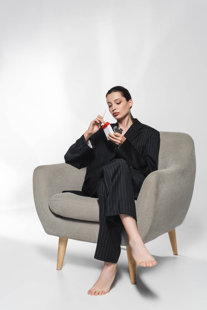 Modelo descalzo en traje que sostiene el cigarrillo y el encendedor mientras está sentado en el sillón sobre fondo gris  - Foto, Imagen