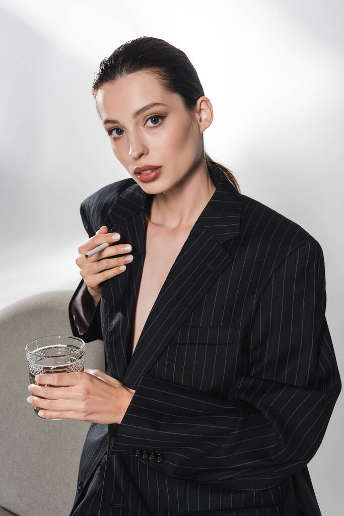 Stílusos nő fekete kabátban kezében üveg whisky és cigaretta közel fotel szürke alapon  - Fotó, kép