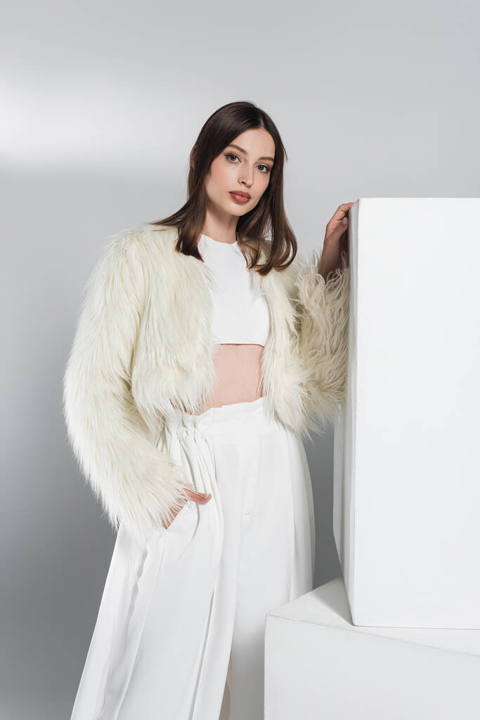 young woman in white faux fur jacket posing with hand in pocket near cubes on grey - Photo, Image