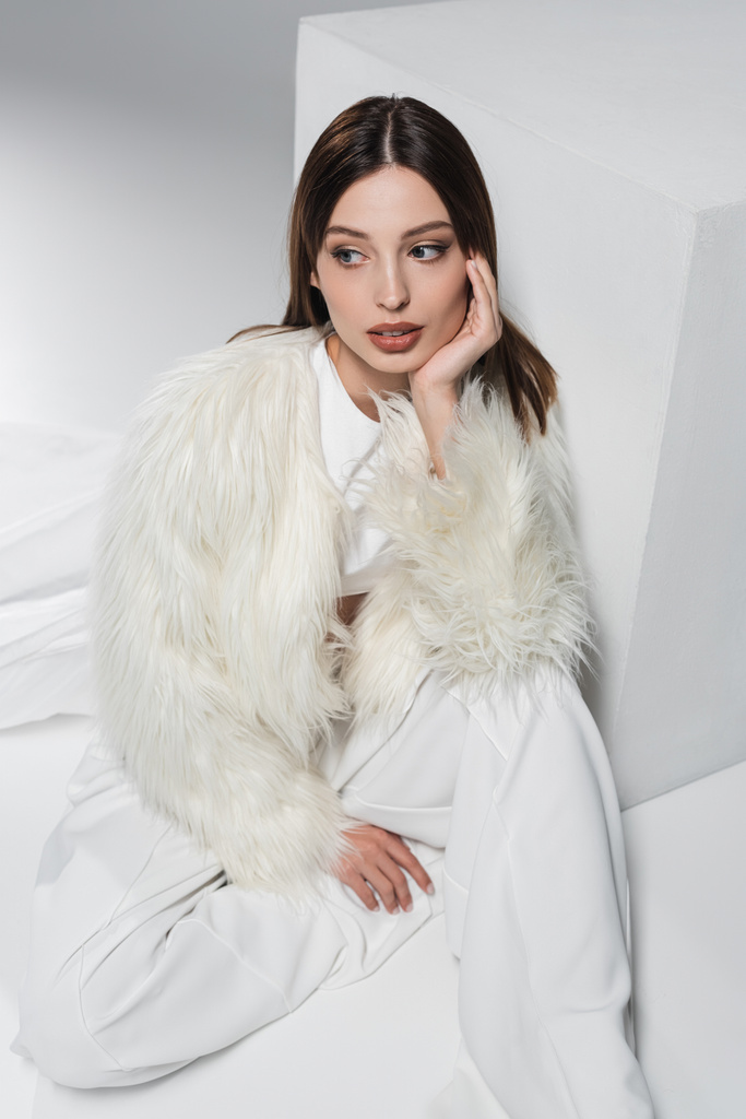 high angle view of young woman in faux fur jacket and total white outfit sitting near cube on grey - Photo, Image