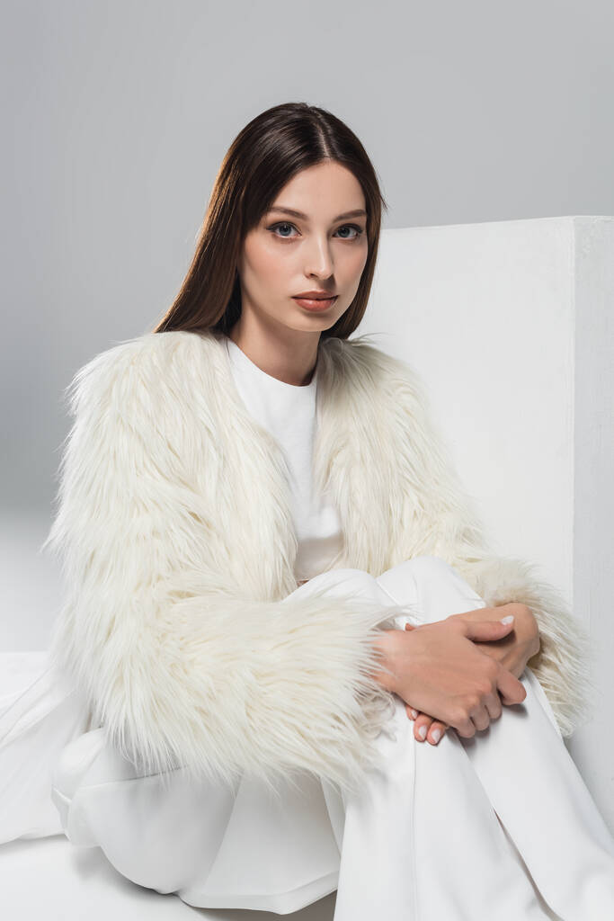 stylish young woman in white faux fur jacket looking at camera near cube on grey - Photo, Image