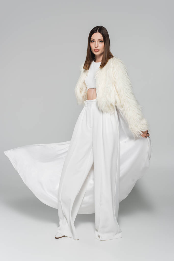 volledige lengte van trendy vrouw in volledig witte outfit en faux fur jas staan op grijs - Foto, afbeelding