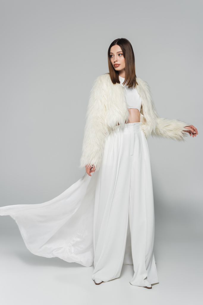 teljes hossza fiatal nő teljesen fehér ruhában és divatos műszőrme kabát pózol szürke - Fotó, kép