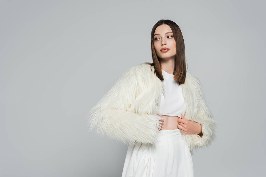 young woman in totally white outfit and trendy faux fur jacket posing isolated on grey - Photo, Image
