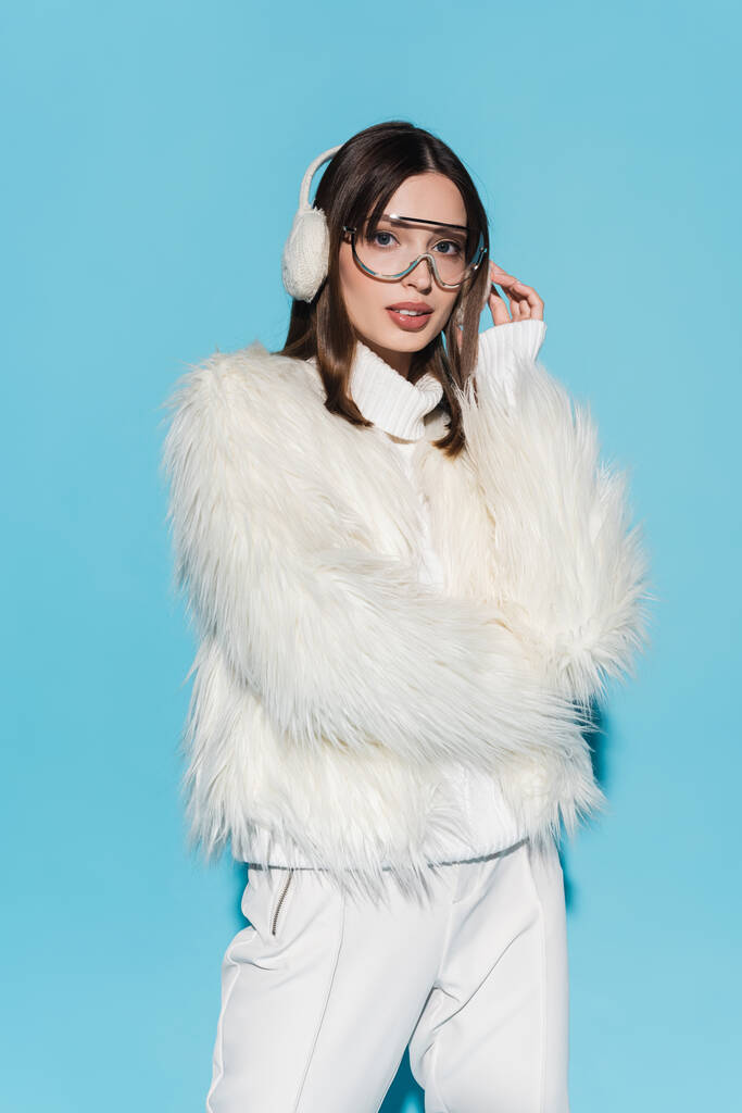 élégant jeune femme en hiver cache-oreilles et lunettes posant dans une veste en fausse fourrure blanche sur bleu - Photo, image