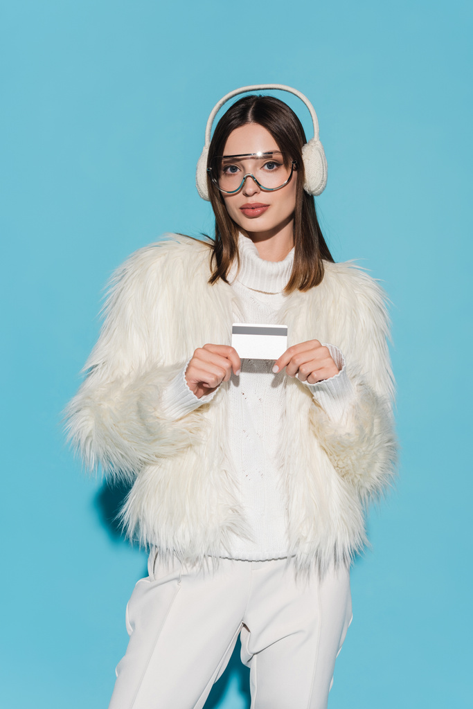 νεαρή γυναίκα με γυαλιά ηλίου και κομψό faux γούνα σακάκι κρατώντας πιστωτική κάρτα σε μπλε - Φωτογραφία, εικόνα