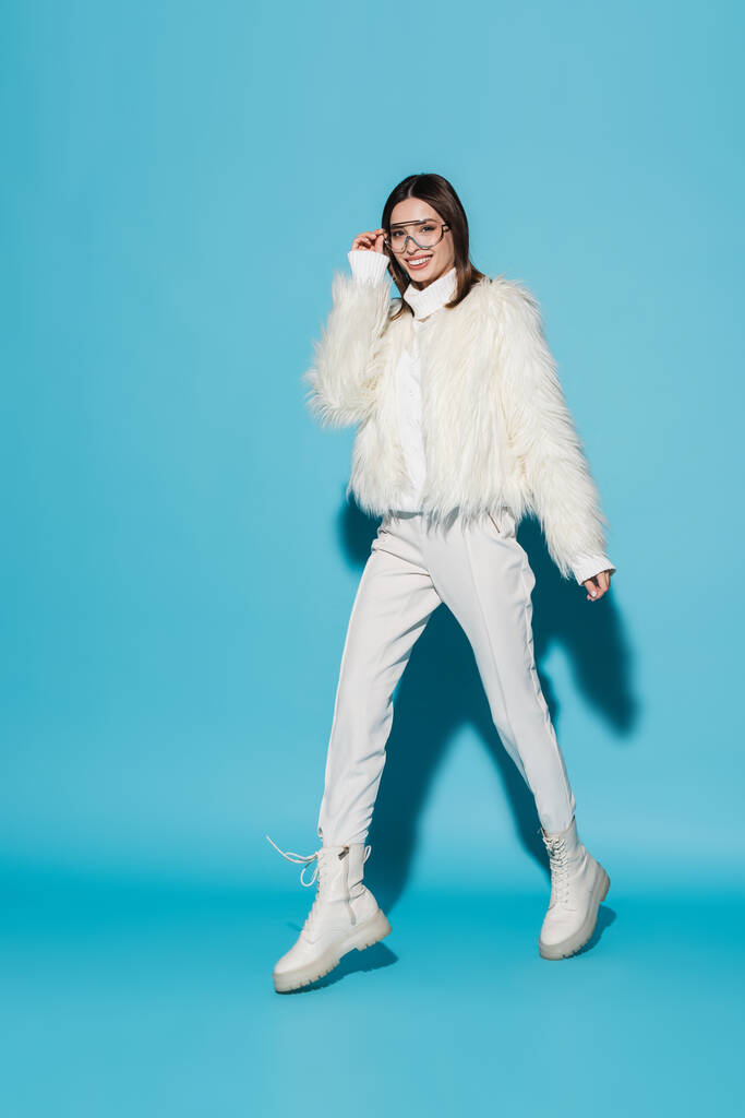 full length of cheerful woman in faux fur jacket and stylish eyeglasses walking on blue - Photo, Image