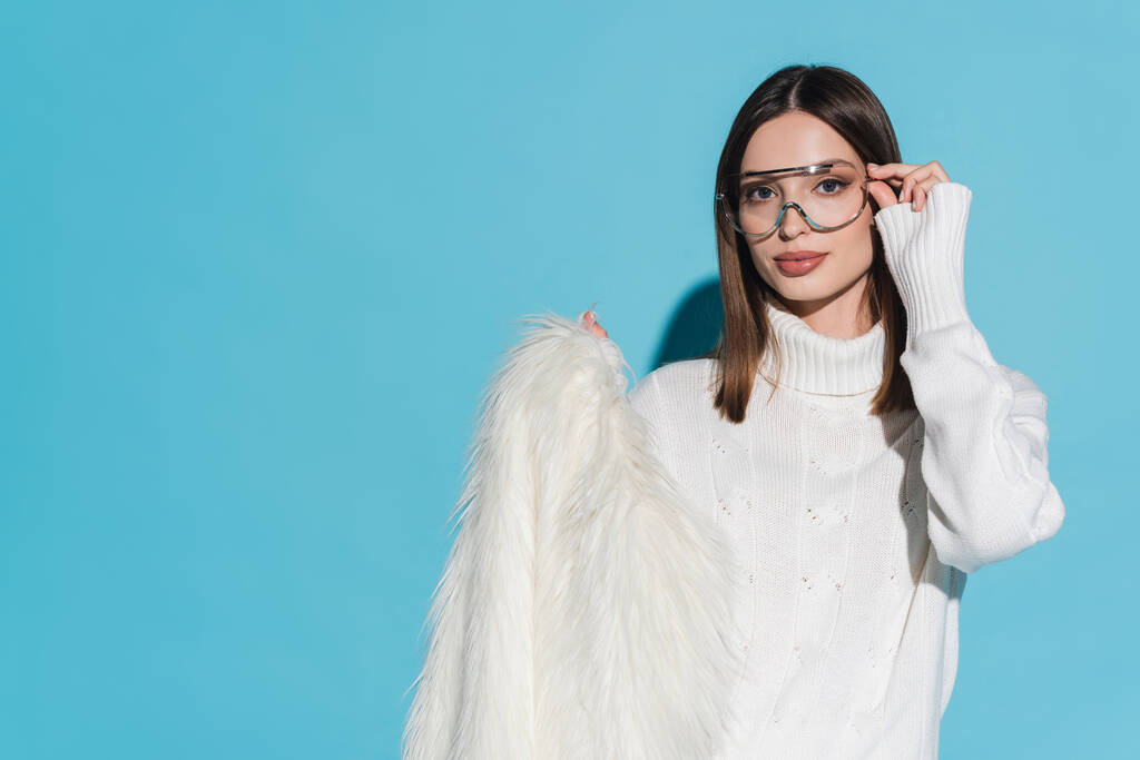 stijlvolle jonge vrouw aanpassen trendy bril en het houden van witte faux fur jas op blauw - Foto, afbeelding