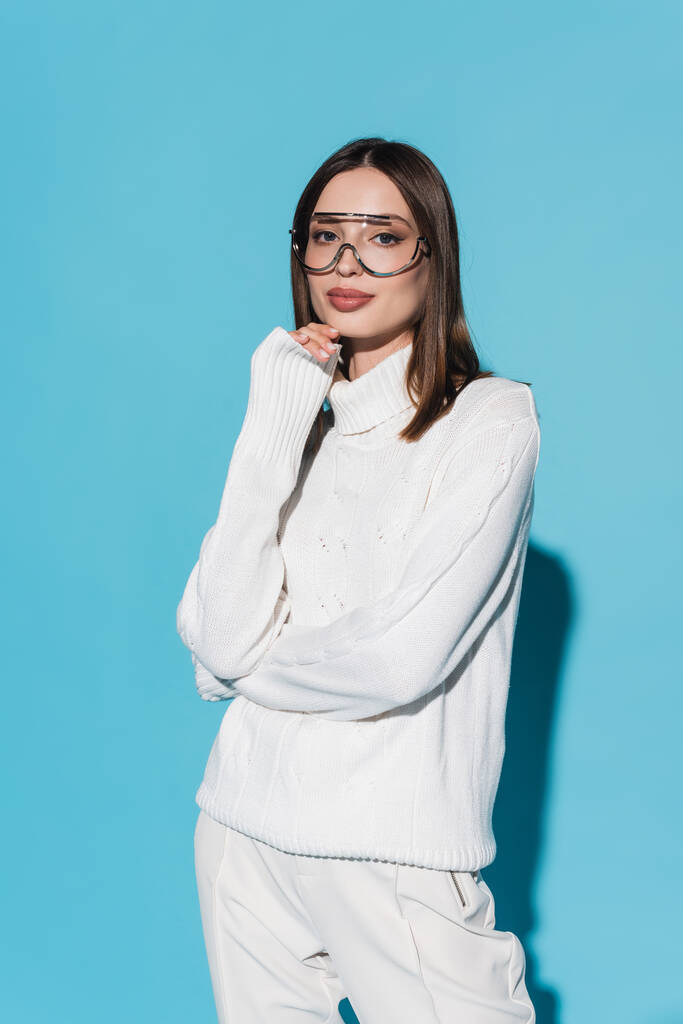 stylische junge Frau mit Brille und weißem Rollkragen blickt in die Kamera auf blauem Grund - Foto, Bild