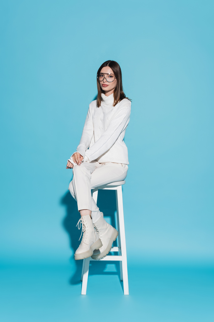 comprimento total de mulher jovem e elegante em roupa branca total sentado em cadeira alta em azul - Foto, Imagem