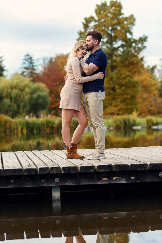 Elegante jovem grávida e seu marido bonito andando de mãos dadas na ponte de madeira através do lago no parque de outono. - Foto, Imagem