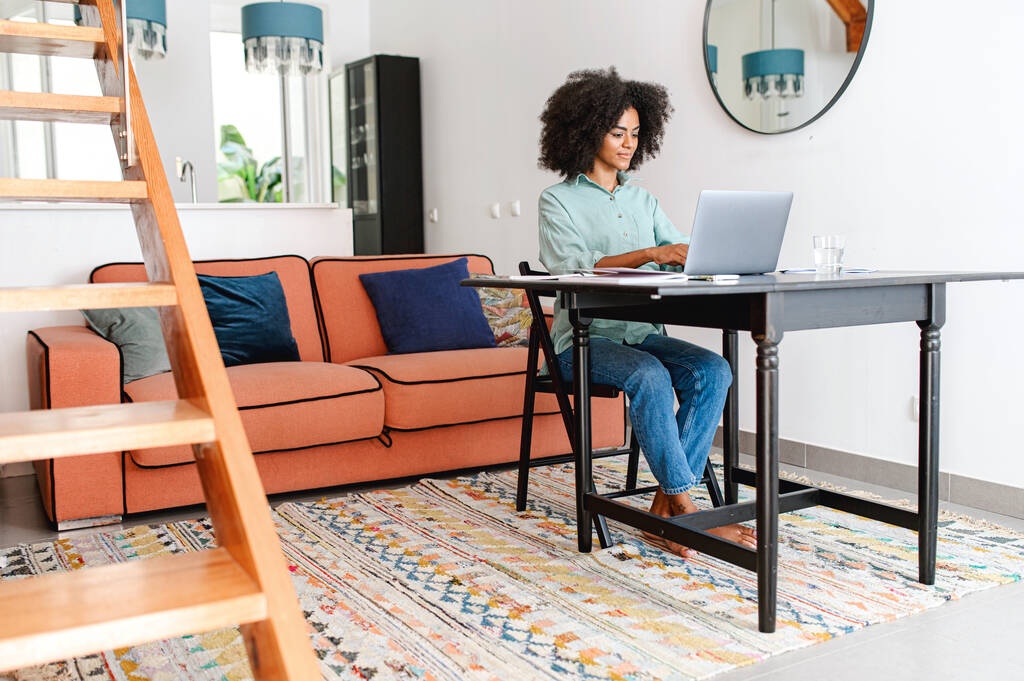 Concentrated young multiracial businesswoman in casual wear sitting at the desk in the home office, working remotely on the laptop. Smiling freelancer woman typing, answering emails - Photo, Image