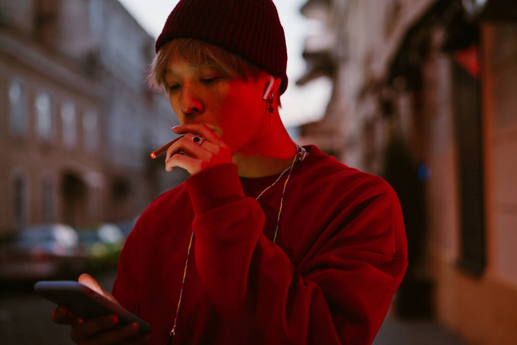 Aasialainen poika kuulokkeissa matkapuhelimella tupakoidessaan kaupungin kadulla - Valokuva, kuva