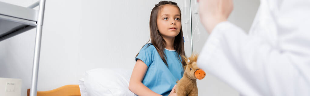 Niño con juguete suave mirando al pediatra borroso en la sala del hospital, pancarta  - Foto, Imagen