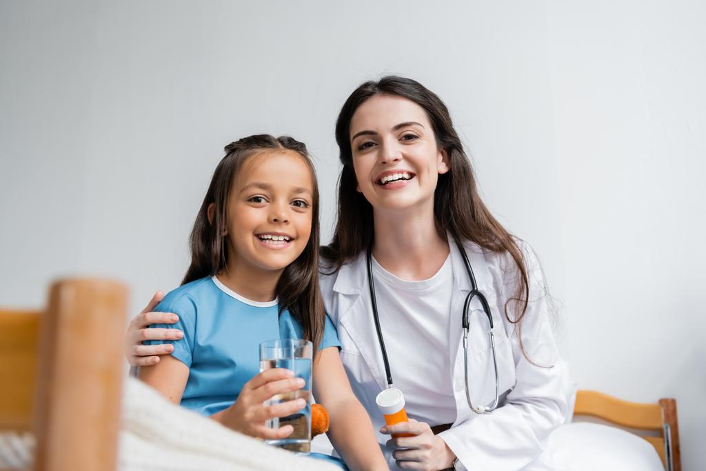 Pediatra sorridente e criança em vestido de paciente segurando vidro de água e pílulas na enfermaria do hospital  - Foto, Imagem