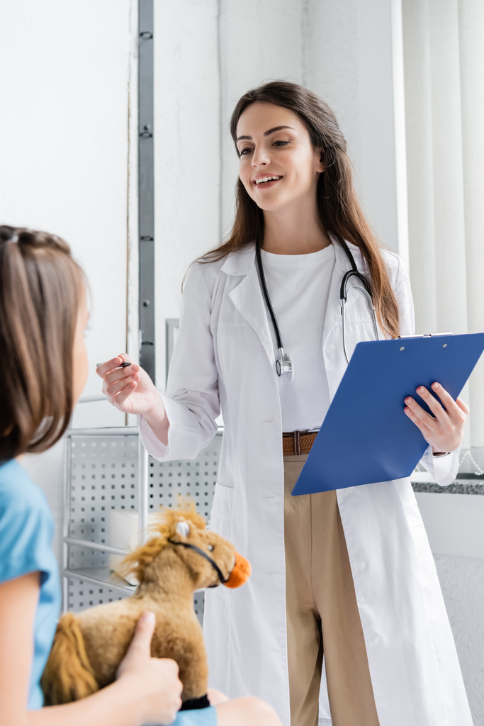 Médico sonriente sosteniendo el portapapeles y hablando con un niño con juguete en la sala del hospital  - Foto, Imagen