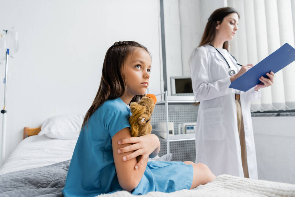Mädchen im Patientenkleid hält Spielzeug in der Nähe des Arztes auf Krankenhausbett  - Foto, Bild