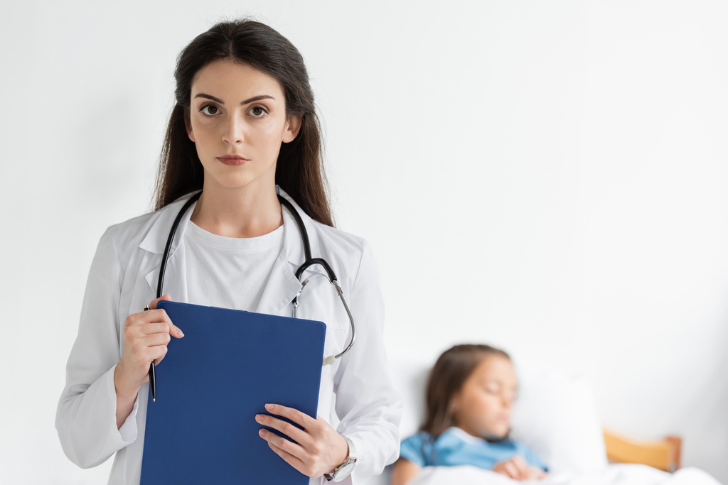Pediatrician holding clipboard and looking at camera near blurred child in clinic  - Photo, Image