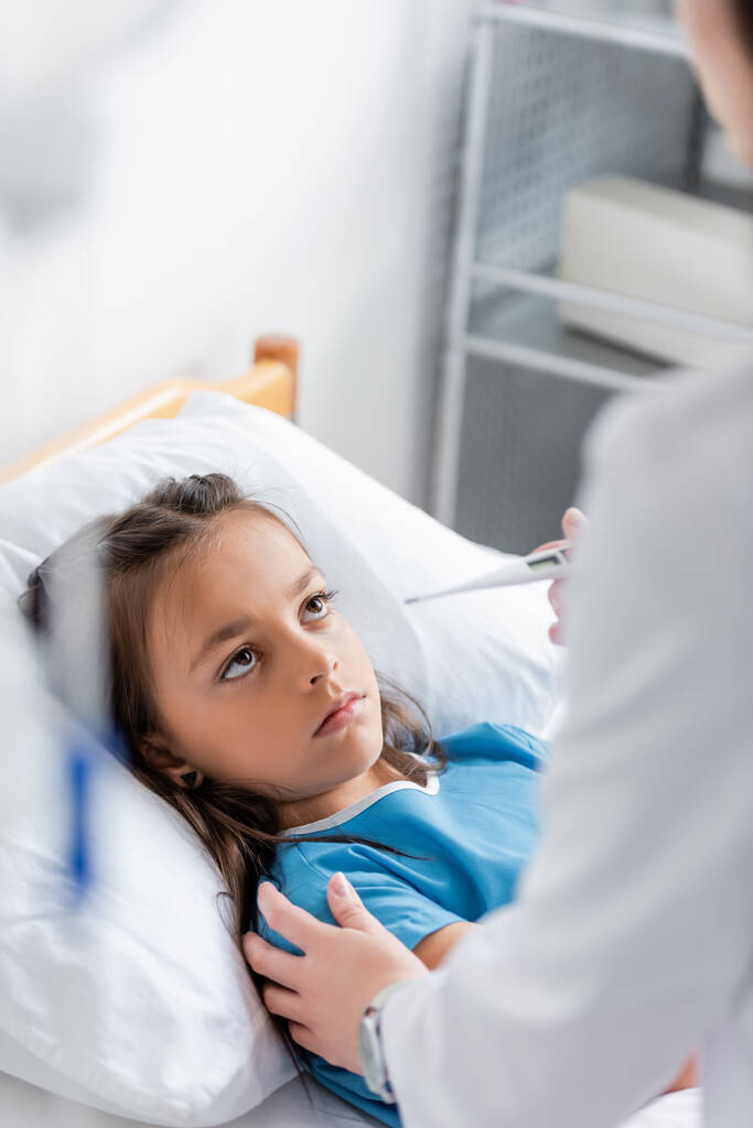 Médecin tenant un thermomètre et touchant l'enfant sur le lit de la clinique  - Photo, image