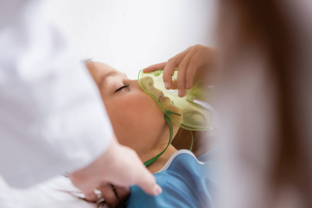 Enfant tenant un masque à oxygène près d'un médecin flou dans la salle d'hôpital  - Photo, image