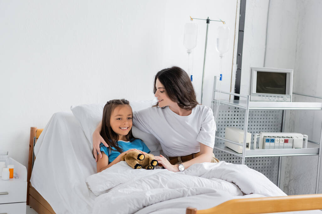 Pozytywne kobieta przytula córkę z zabawką na łóżku kliniki  - Zdjęcie, obraz