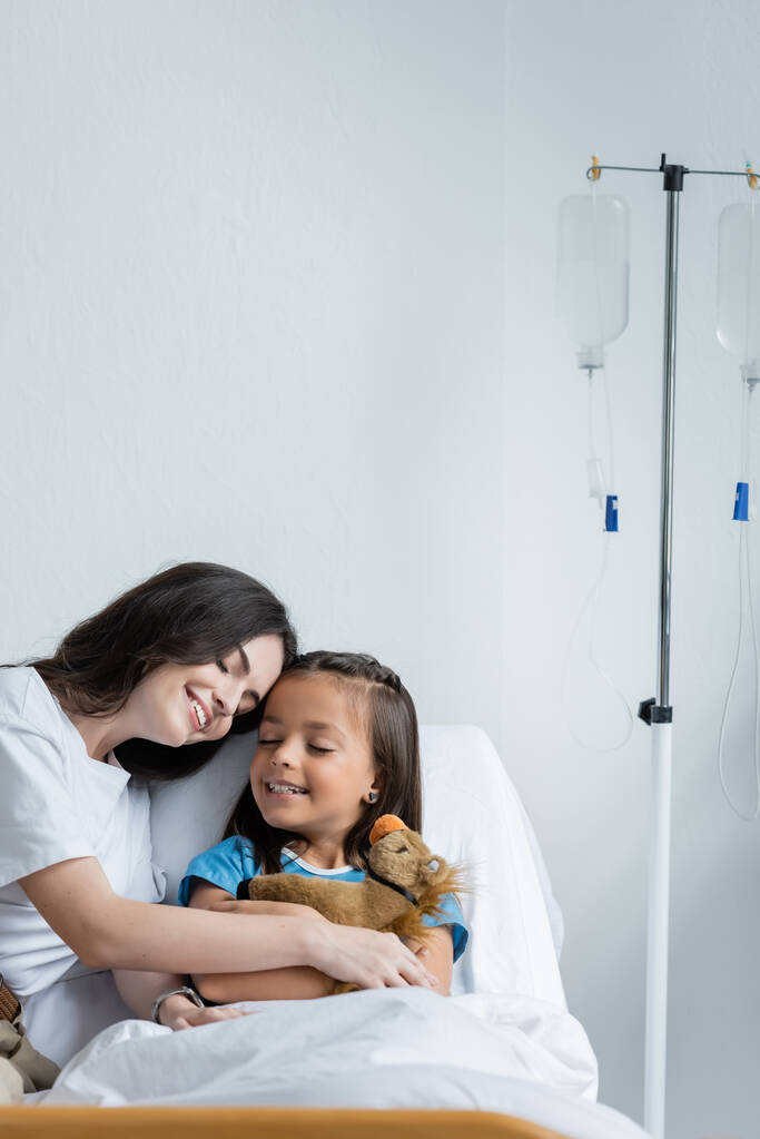 Parent touching hand of smiling child with soft toy on bed in clinic  - Photo, Image