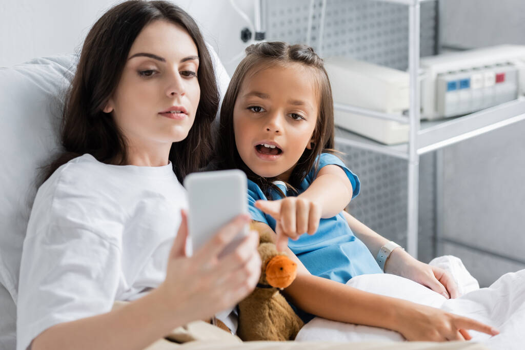 Kind zeigt in der Nähe von Mama auf Smartphone und Kuscheltier auf Bett in Klinik  - Foto, Bild