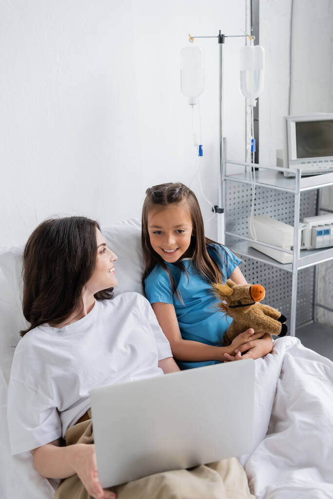 Sonriente niño en vestido de paciente mirando a la computadora portátil cerca de mamá en la cama del hospital  - Foto, Imagen