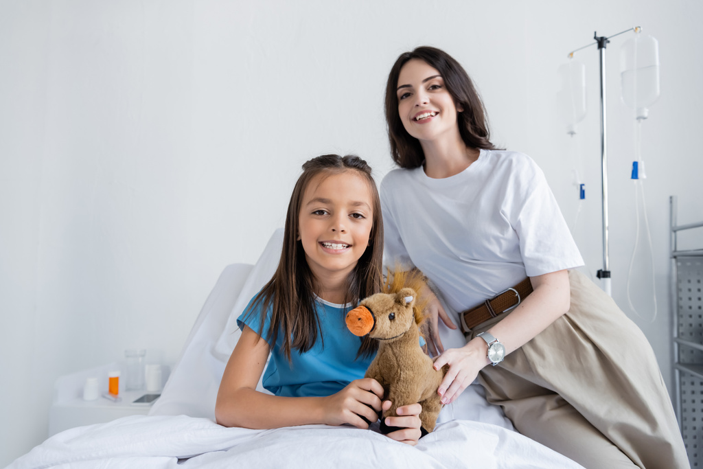 Lächelndes Kind im Patientenkleid hält Kuscheltier in der Nähe der Mutter im Krankenhaus  - Foto, Bild