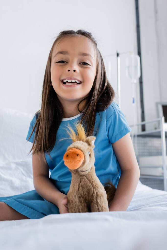 Улыбающийся ребенок в больничном халате смотрит в камеру возле мягкой игрушки на кровати в клинике  - Фото, изображение