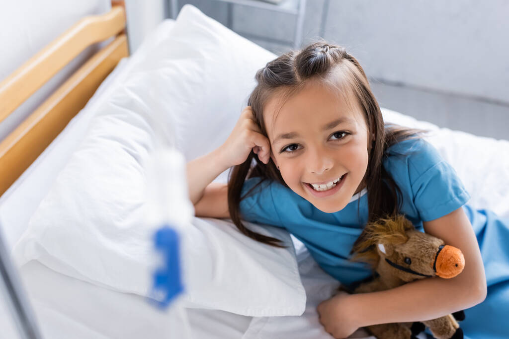 Hochwinkelaufnahme des fröhlichen Mädchens, das Spielzeug auf dem Bett in der Klinik hält  - Foto, Bild