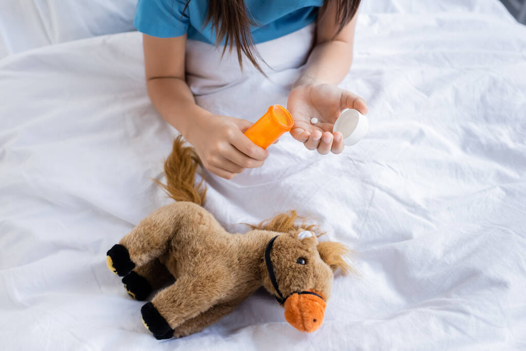 Vista cortada de crianças segurando pílulas perto de brinquedo na cama na clínica  - Foto, Imagem